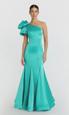 A woman stands against a white background wearing the Mac Duggal 68703 long formal dress in teal, featuring a fitted, floor-length satin design with an asymmetrical neckline and a statement ruffle on one shoulder.