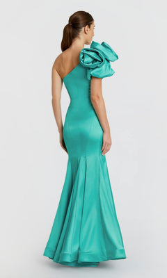 A woman stands facing away against a plain white background, wearing the Mac Duggal 68703 long formal dress—a fitted, teal, one-shoulder satin gown featuring a large ruffled shoulder detail.