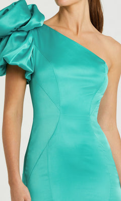 A woman models the Mac Duggal 68703 long formal dress—a fitted turquoise satin gown with a one-shoulder design and dramatic ruffled left sleeve—posed against a plain background.
