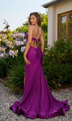 A woman in the Cut-Out Strapless Long Prom Dress: Atria 6870H stands outdoors on a stone path, surrounded by greenery and flowers, glancing over her shoulder.