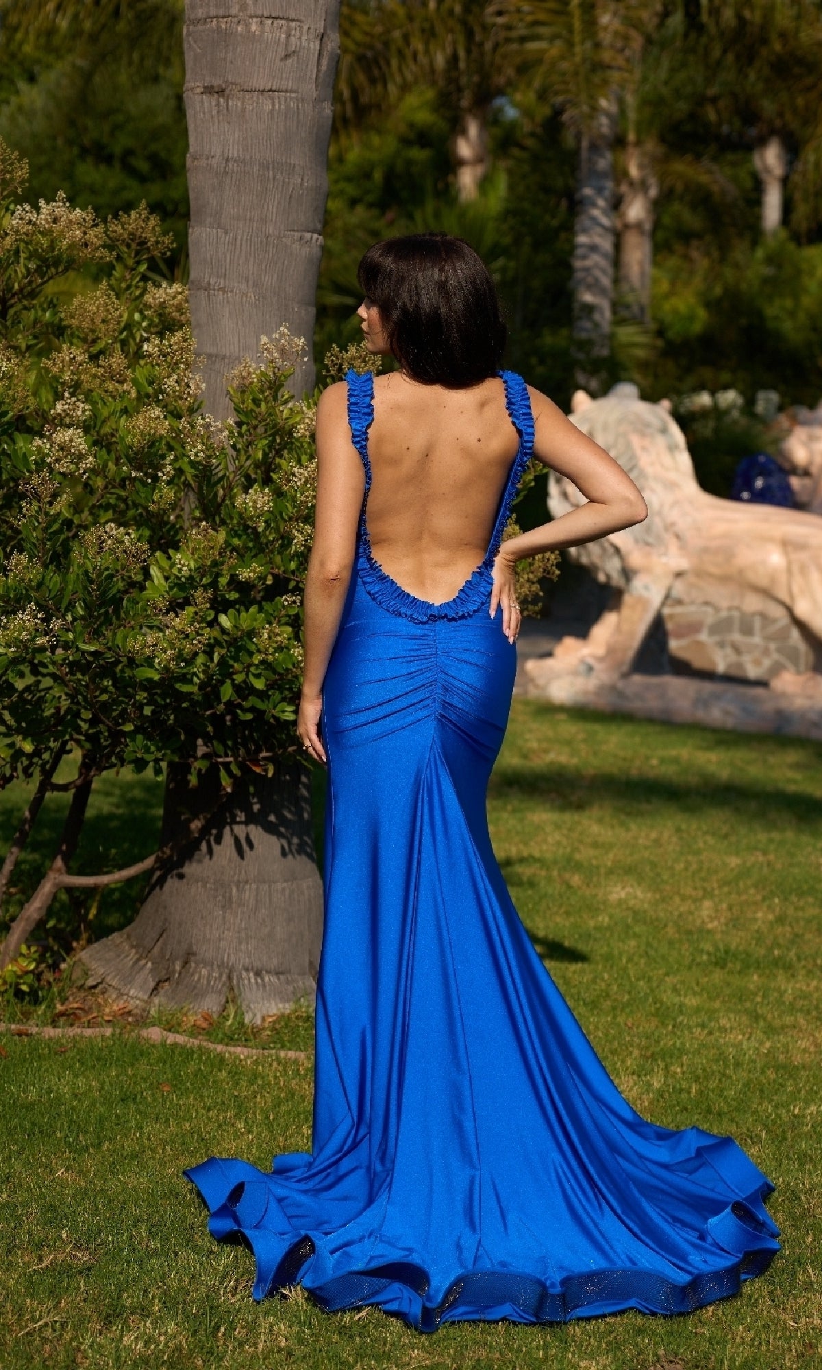 A woman stands outdoors on grass, facing away, wearing the Atria 6871H Ruffle Open-Back Long Prom Dress. Trees and greenery can be seen in the background.