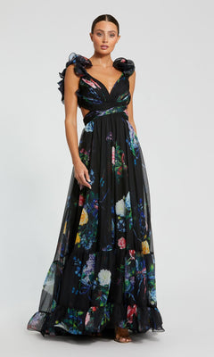 A woman wears the Mac Duggal 68746 Black Floral Formal Evening Gown, featuring ruffled shoulders, side cutouts, and a floor-length skirt, standing against a plain light background.