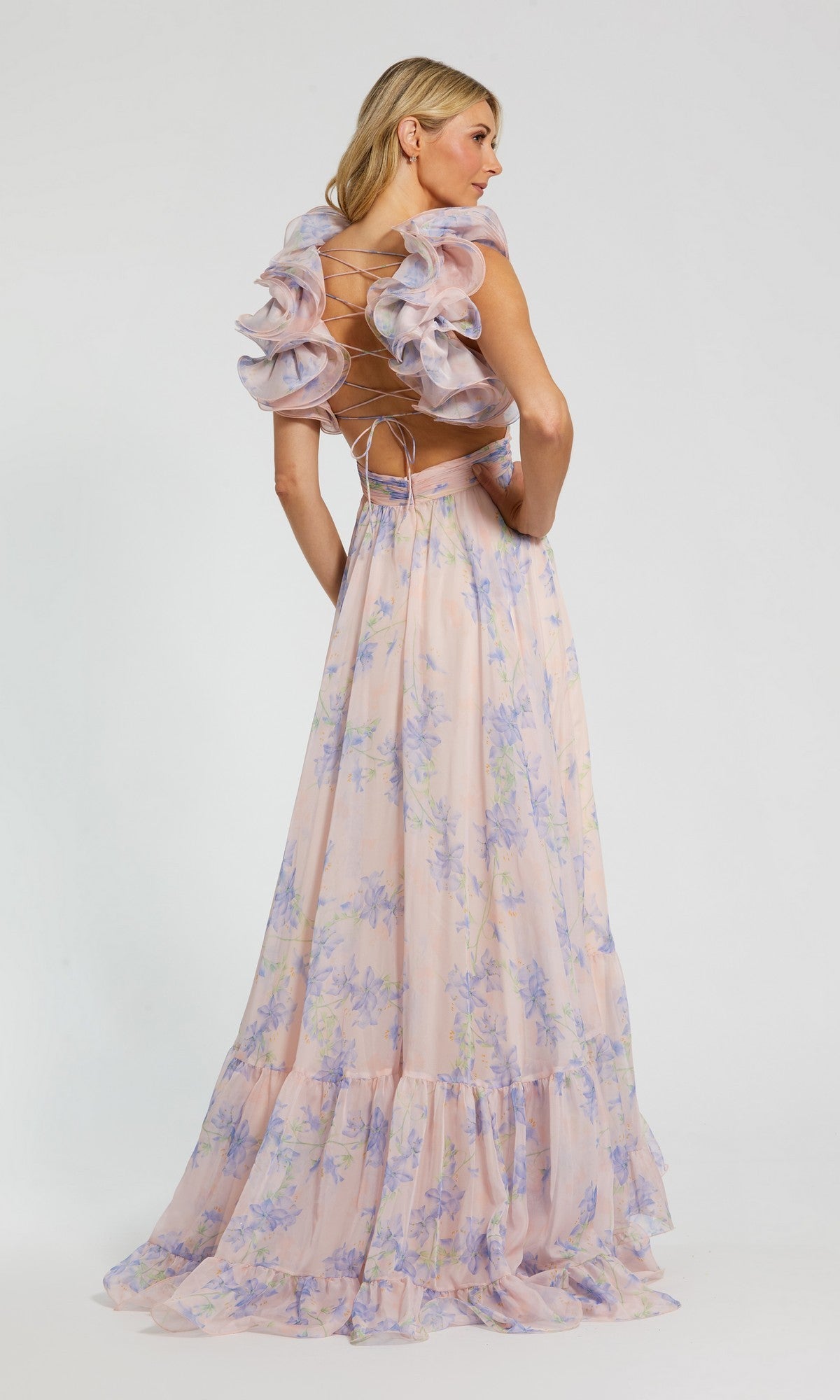 A woman in the Mac Duggal 68753 Long Formal Dress—a violet floral chiffon gown with ruffled sleeves and an open back—stands sideways, looking over her shoulder against a plain background.