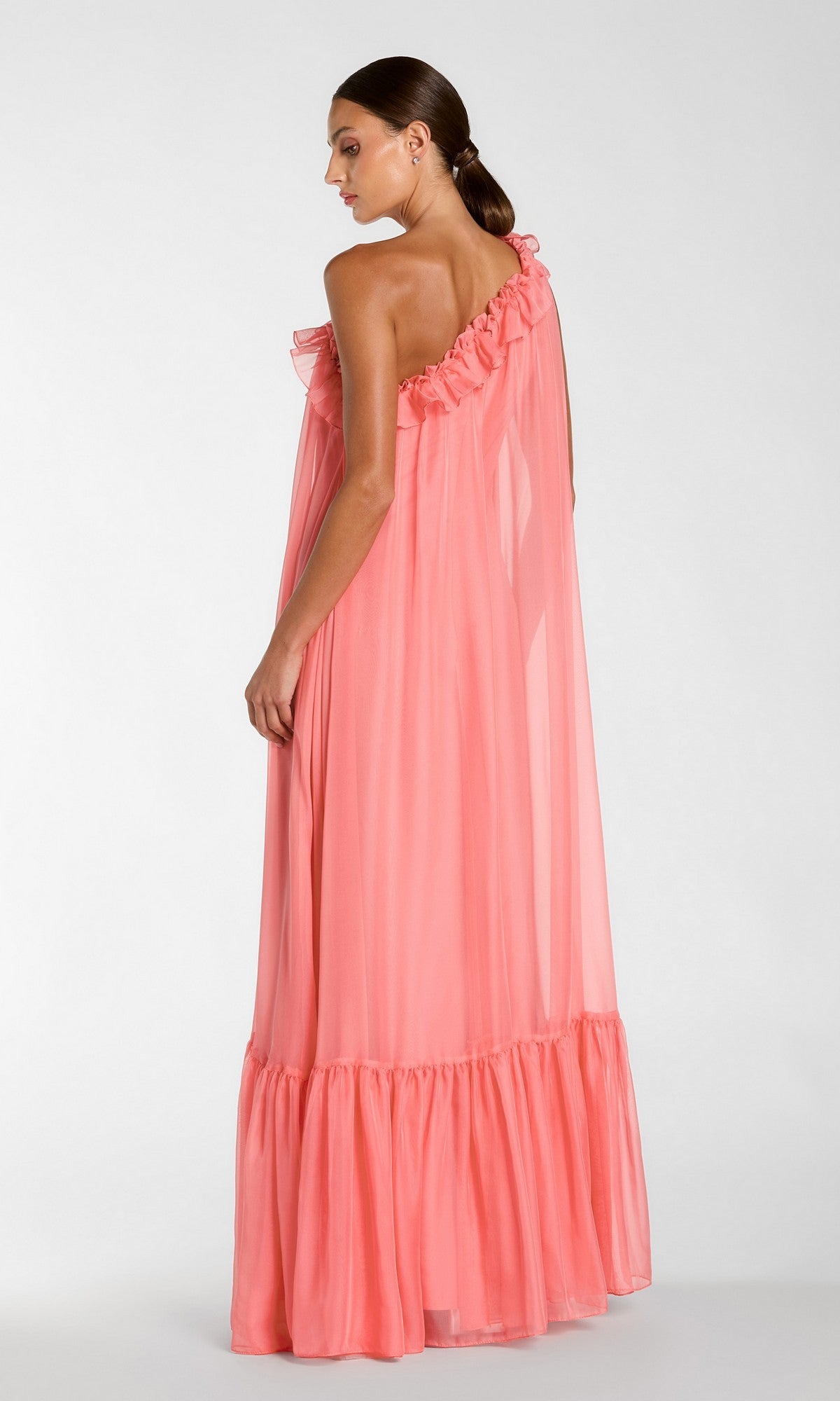 A woman with brown hair in a low ponytail models the Mac Duggal 68757 Long Formal Dress—a coral pink, one-shoulder chiffon gown featuring elegant ruffles and a flowing maxi silhouette. She stands facing away, looking over her right shoulder against a plain background.