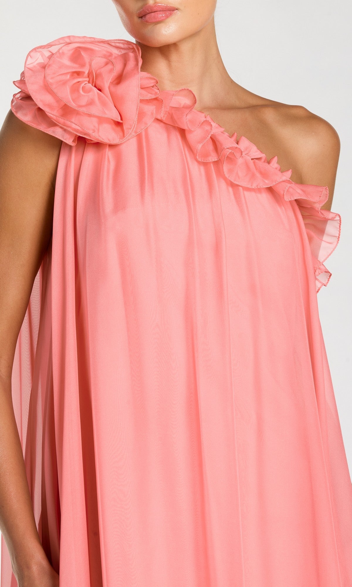 A person wearing the Mac Duggal 68757 long formal dress, featuring a pink, one-shoulder chiffon design with ruffle details and a large fabric flower accenting the shoulder.