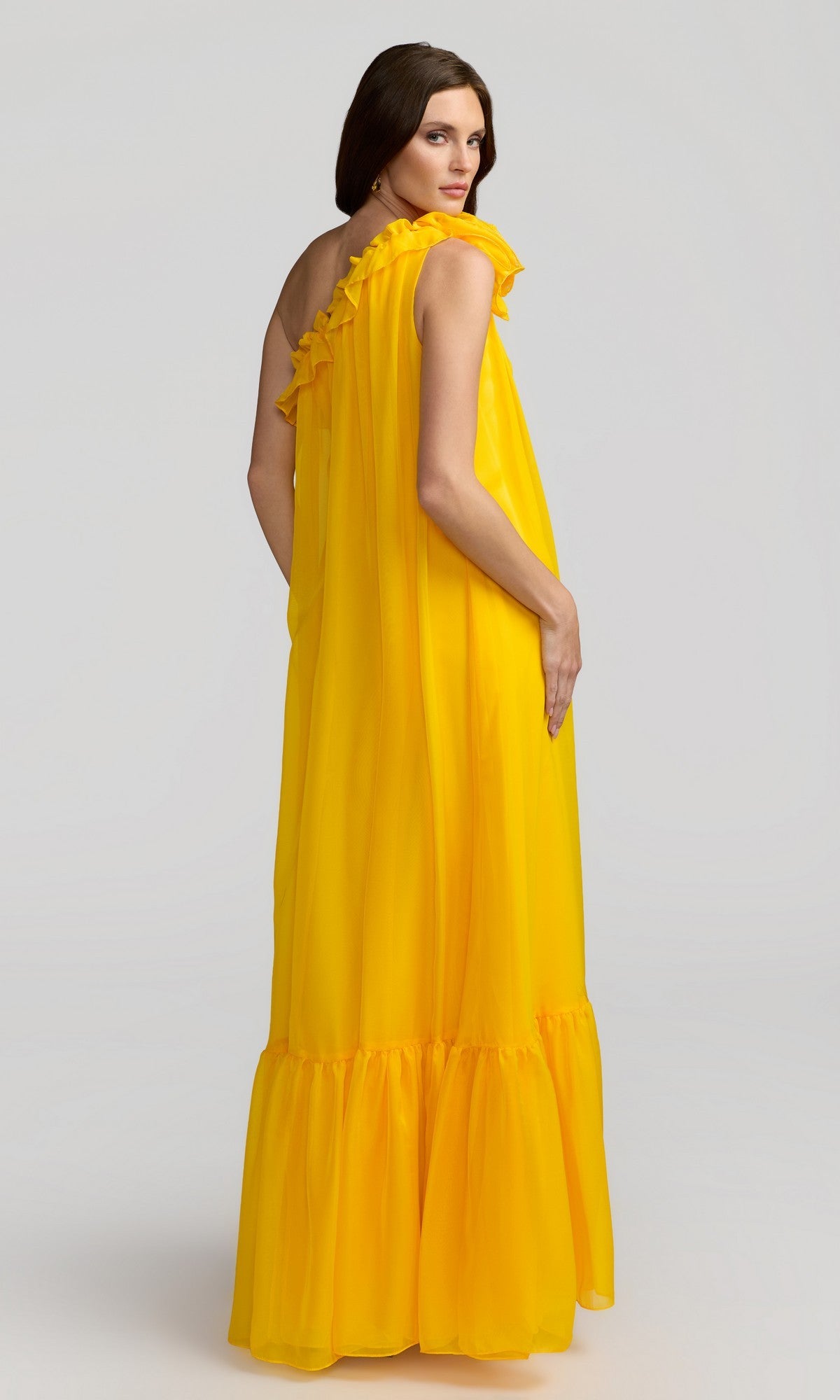 A woman poses against a plain light background, turning her head over her shoulder while wearing the Mac Duggal 68757 long formal dress in bright yellow with ruffled details.