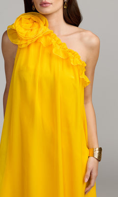 A woman wearing the Long Formal Dress: Mac Duggal 68757 in bright yellow, featuring a one-shoulder chiffon design with ruffle detailing and a large floral accent, accessorized with a gold cuff bracelet.