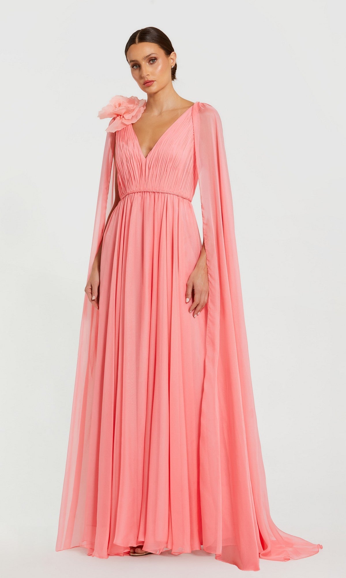 A woman stands against a plain white background wearing the Mac Duggal 68768 long formal dress in vibrant hibiscus, featuring flowing chiffon fabric, elegant cape sleeves, and a dramatic oversized flower accent on one shoulder.