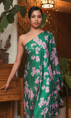 A woman stands indoors by a piano, wearing the Mac Duggal 68787 long green print formal dress. Pink floral details highlight the one-shoulder style. Plants and a beaded curtain decorate the background.