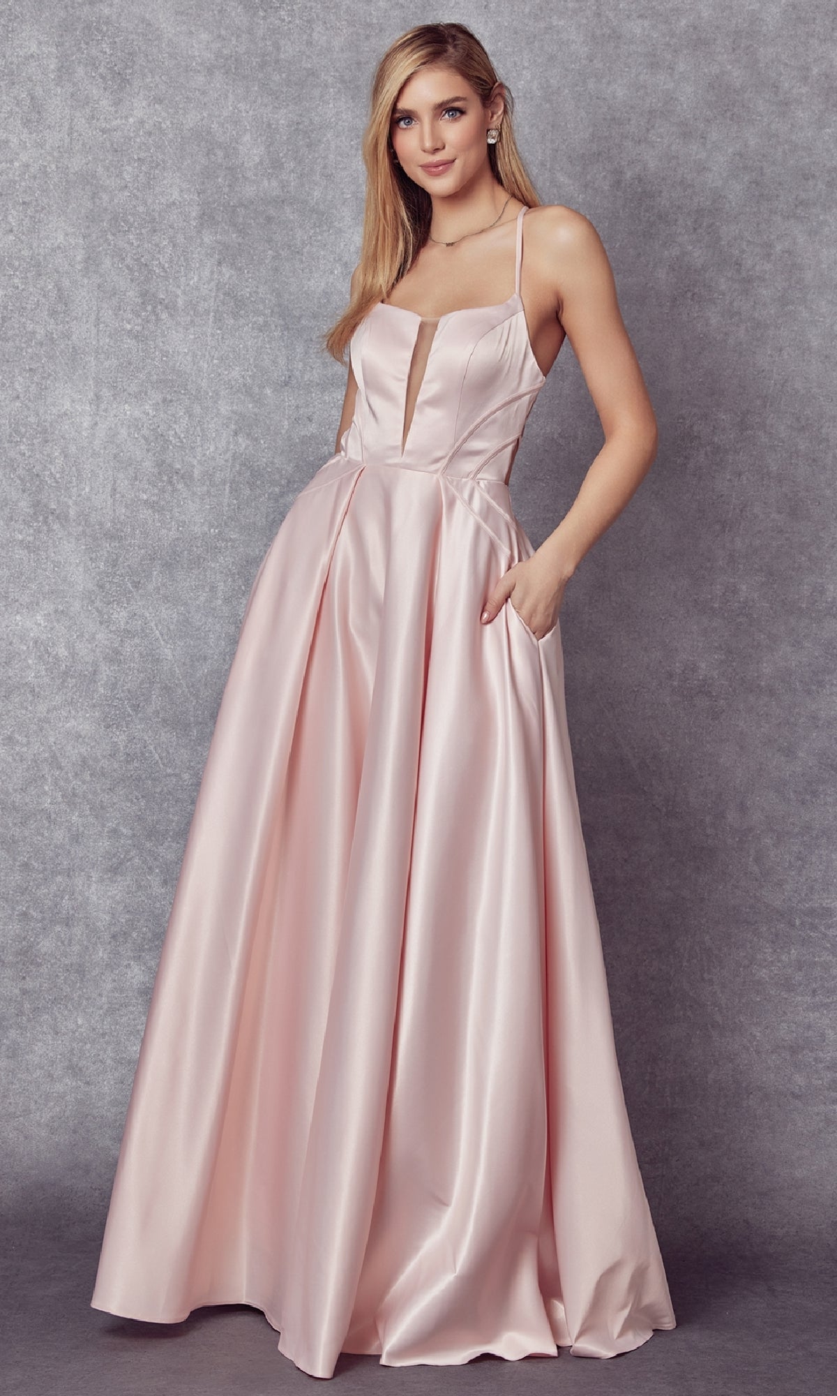 A woman stands against a gray textured background wearing the Juliet 687 long prom dress, featuring an open back, sheer V-neckline, sleeveless pale pink satin, fitted bodice, and full skirt.
