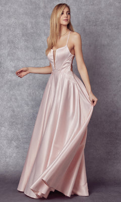 A woman in the Long Prom Dress: Juliet 687—a light pink satin gown with a sheer v-neckline and thin straps—stands against a textured gray background, holding out part of the skirt.