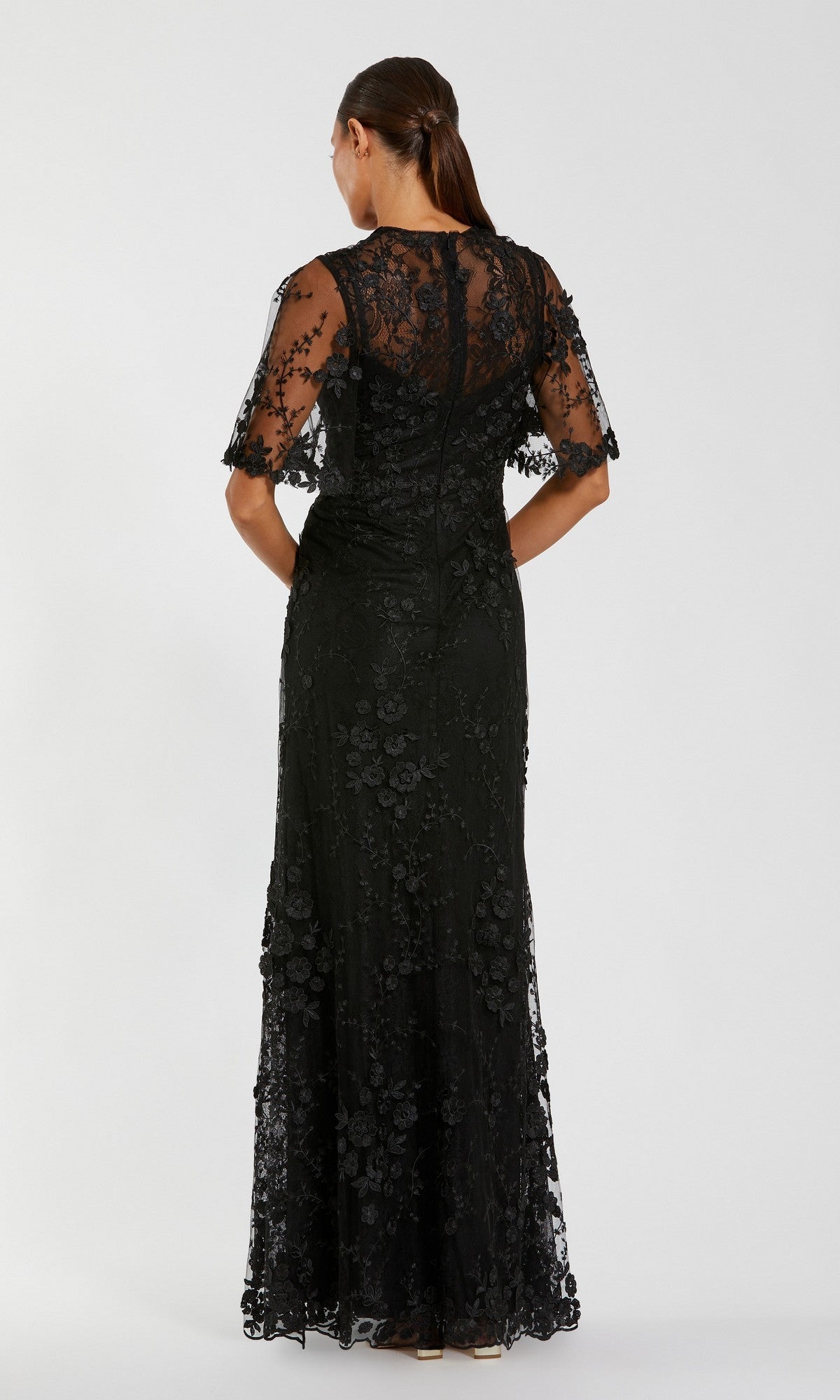 A woman stands facing away, wearing the Mac Duggal 68810 Flutter-Sleeve Long Formal Dress—a floor-length black gown featuring sheer short sleeves and elegant floral lace details.