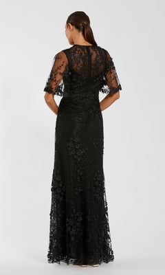 A woman stands facing away, wearing the Mac Duggal 68810 Flutter-Sleeve Long Formal Dress—a floor-length black gown featuring sheer short sleeves and elegant floral lace details.