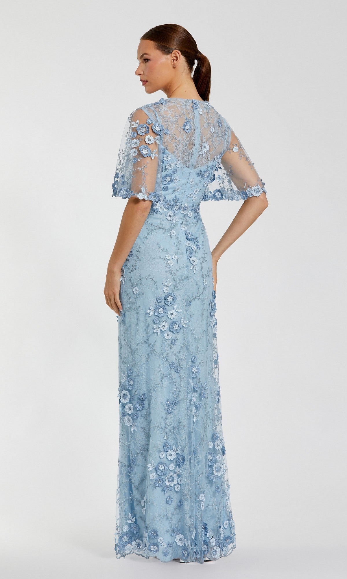 A woman models the Mac Duggal 68810 Flutter-Sleeve Long Formal Dress in light blue lace with floral appliqué and short sleeves, shown from the back against a plain background.