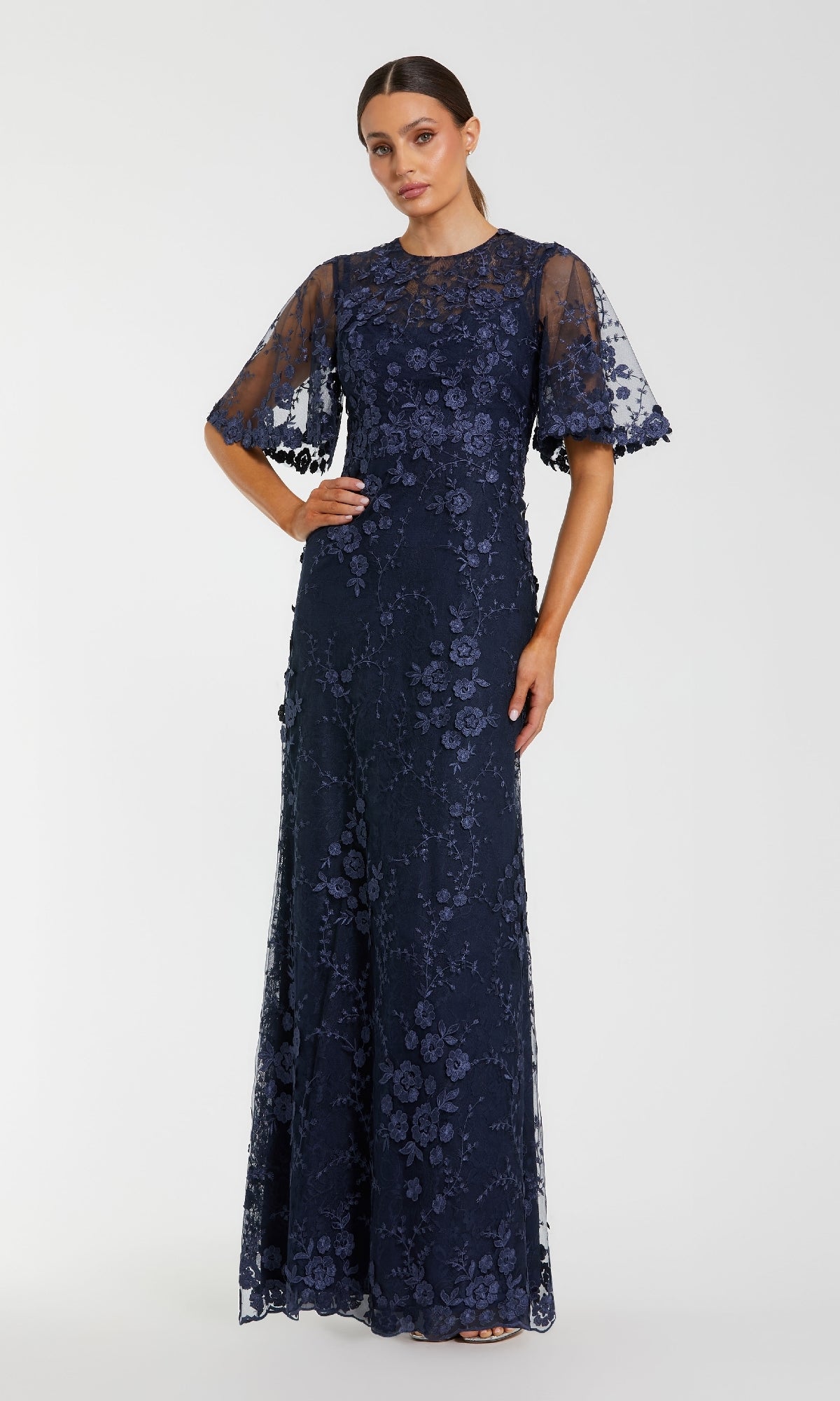 A woman stands against a plain light background in the Mac Duggal 68810 Flutter-Sleeve Long Formal Dress, a navy blue lace gown with floral patterns and sheer, elbow-length sleeves—perfect for the mother of the bride.