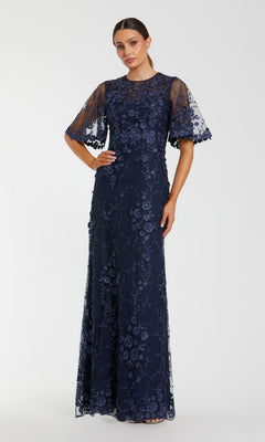 A woman stands against a plain light background in the Mac Duggal 68810 Flutter-Sleeve Long Formal Dress, a navy blue lace gown with floral patterns and sheer, elbow-length sleeves—perfect for the mother of the bride.