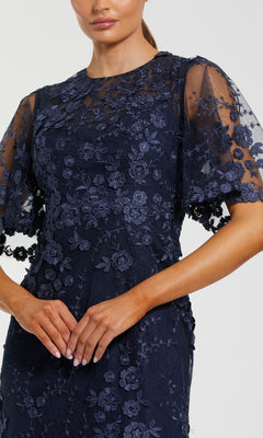 A woman stands with hands clasped, wearing the Mac Duggal 68810 Flutter-Sleeve Long Formal Dress in navy blue lace with floral embroidery and sheer short flutter sleeves.