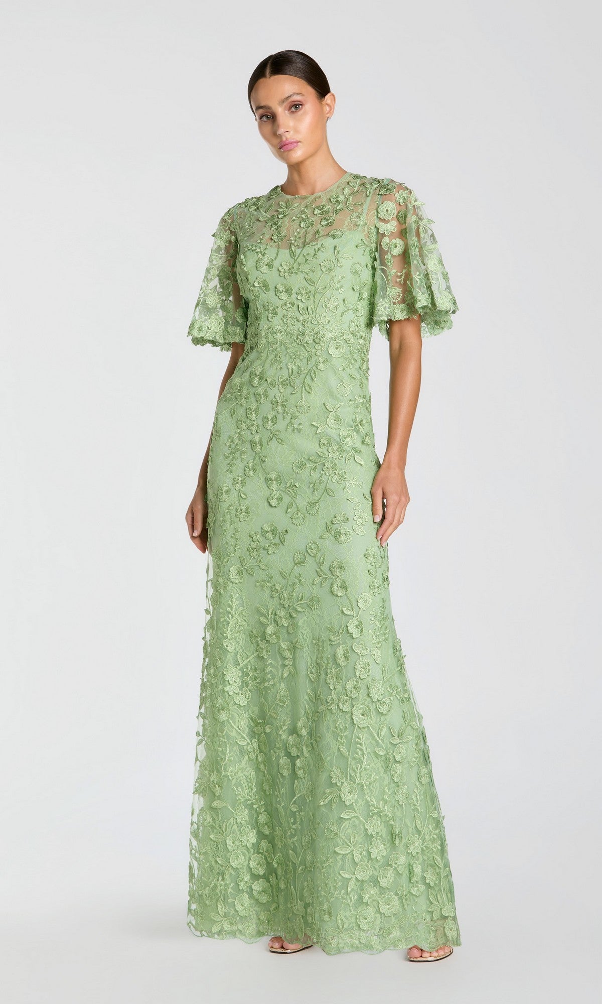 A woman models the Mac Duggal 68810 Flutter-Sleeve Long Formal Dress—a light green lace gown with short flutter sleeves and floral embroidery—posed against a plain, light background.
