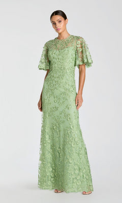 A woman models the Mac Duggal 68810 Flutter-Sleeve Long Formal Dress—a light green lace gown with short flutter sleeves and floral embroidery—posed against a plain, light background.