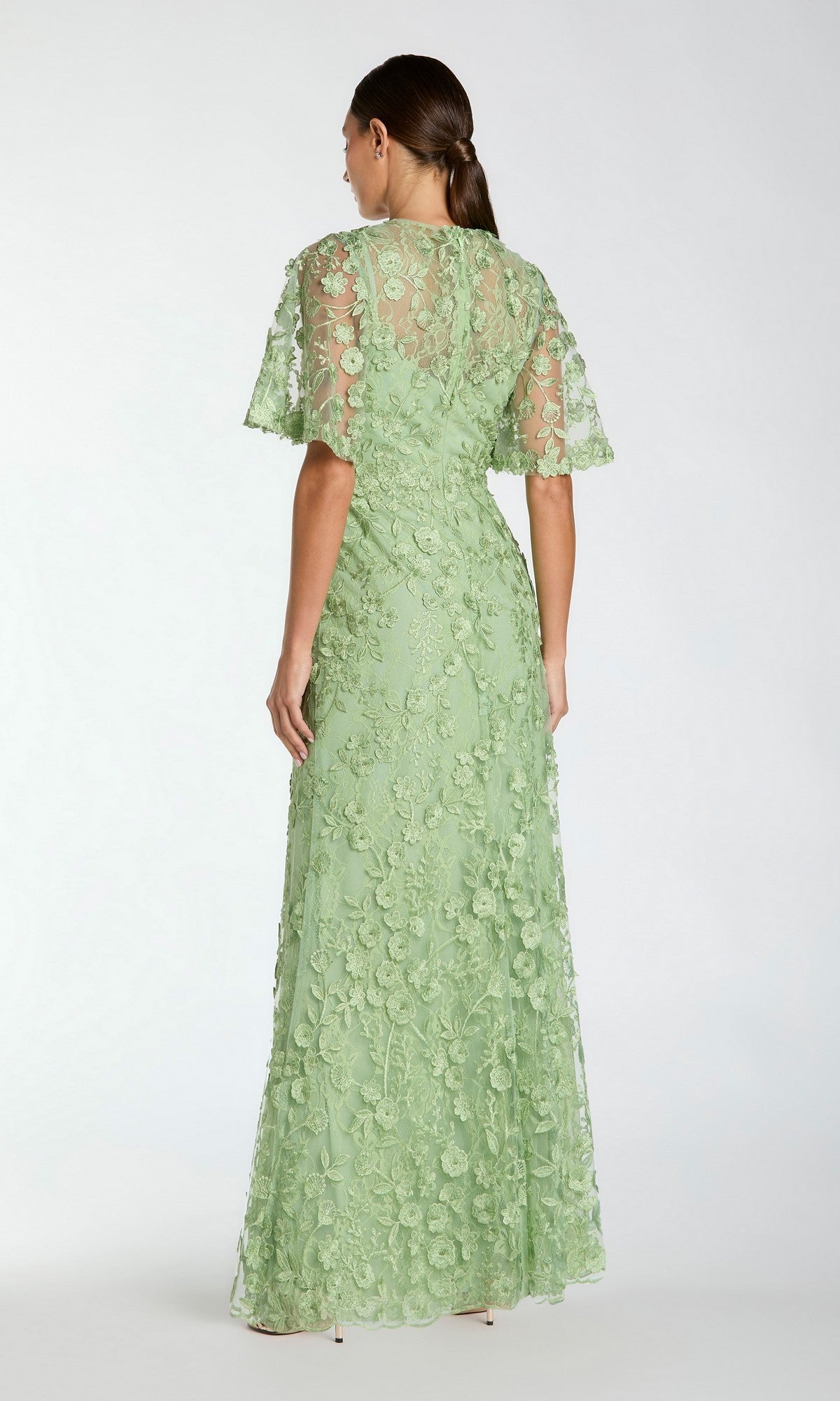 A woman stands with her back to the camera, wearing the Mac Duggal 68810 Flutter-Sleeve Long Formal Dress in light green, featuring floral lace detailing and short flutter sleeves.
