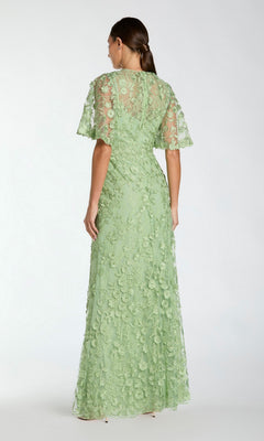 A woman stands with her back to the camera, wearing the Mac Duggal 68810 Flutter-Sleeve Long Formal Dress in light green, featuring floral lace detailing and short flutter sleeves.
