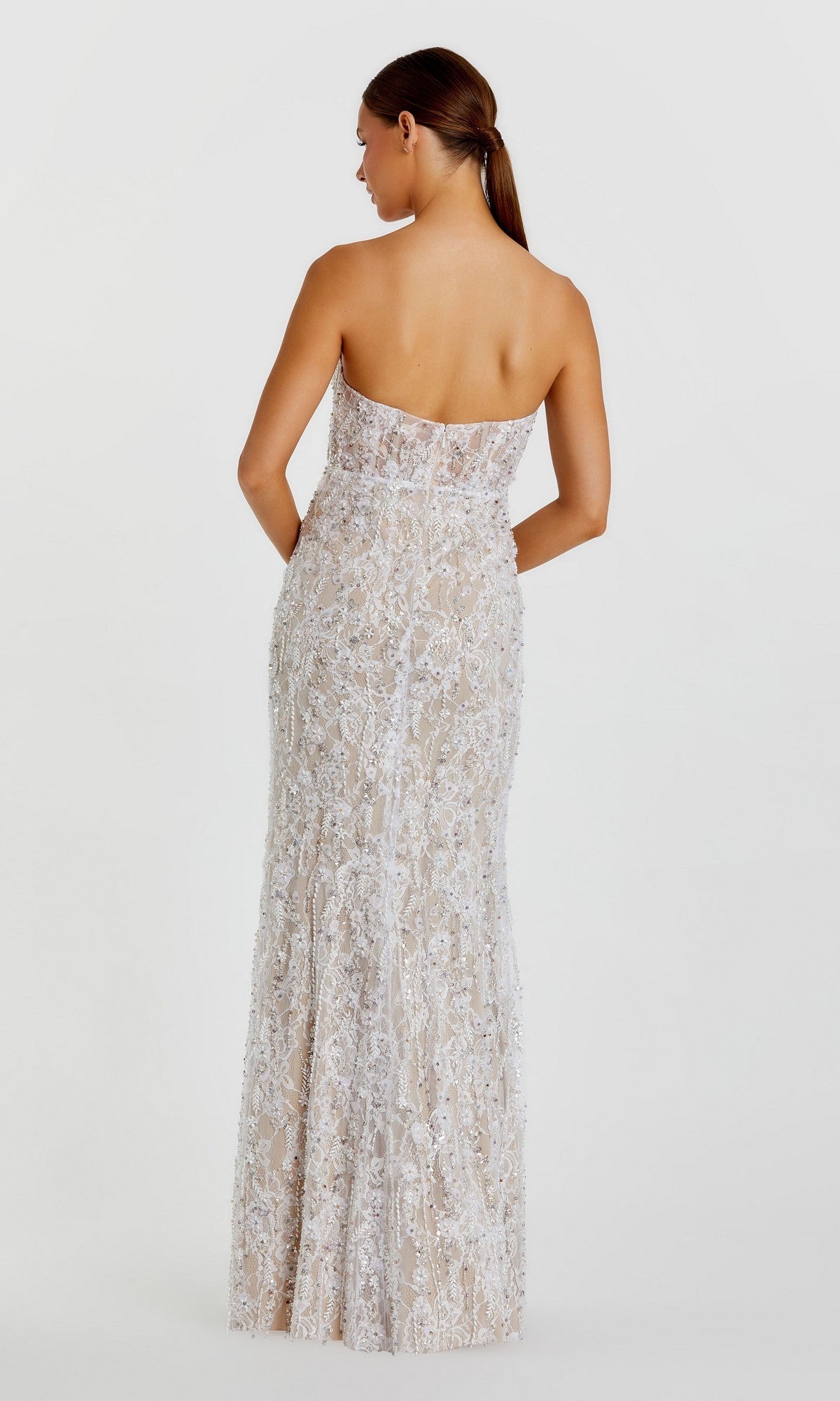 A woman stands facing away against a plain white background, wearing the Long Formal Dress: Mac Duggal 68893—a floor-length, form-fitting strapless gown featuring intricate white lace, shimmering sequin patterns, and delicate floral beadwork.