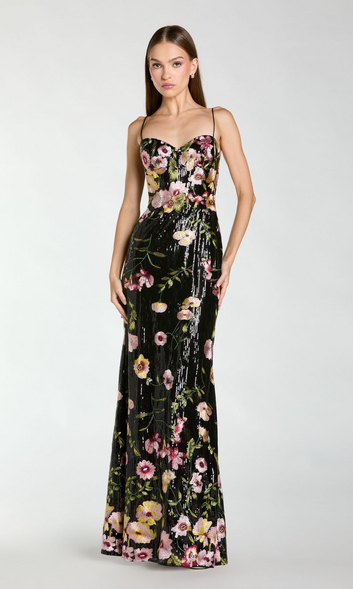 A woman wears the Mac Duggal 68896 long formal dress in Black Multi, featuring thin straps and a vibrant floral pattern, standing against a plain light background.