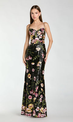 A woman wears the Mac Duggal 68896 long formal dress in Black Multi, featuring thin straps and a vibrant floral pattern, standing against a plain light background.