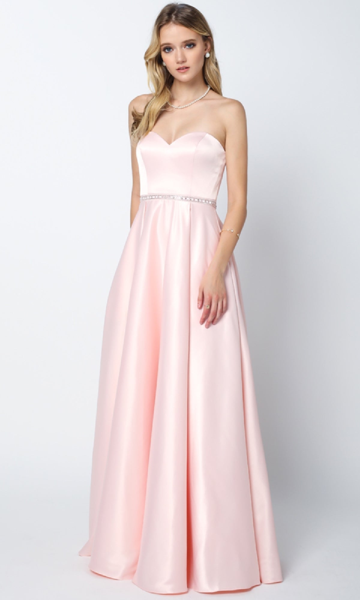 A woman stands against a plain background wearing the Juliet 688 Long Prom Dress—a floor-length, light pink satin gown with a strapless, fitted bodice, subtle waistband embellishment, and a gentle sweetheart neckline.