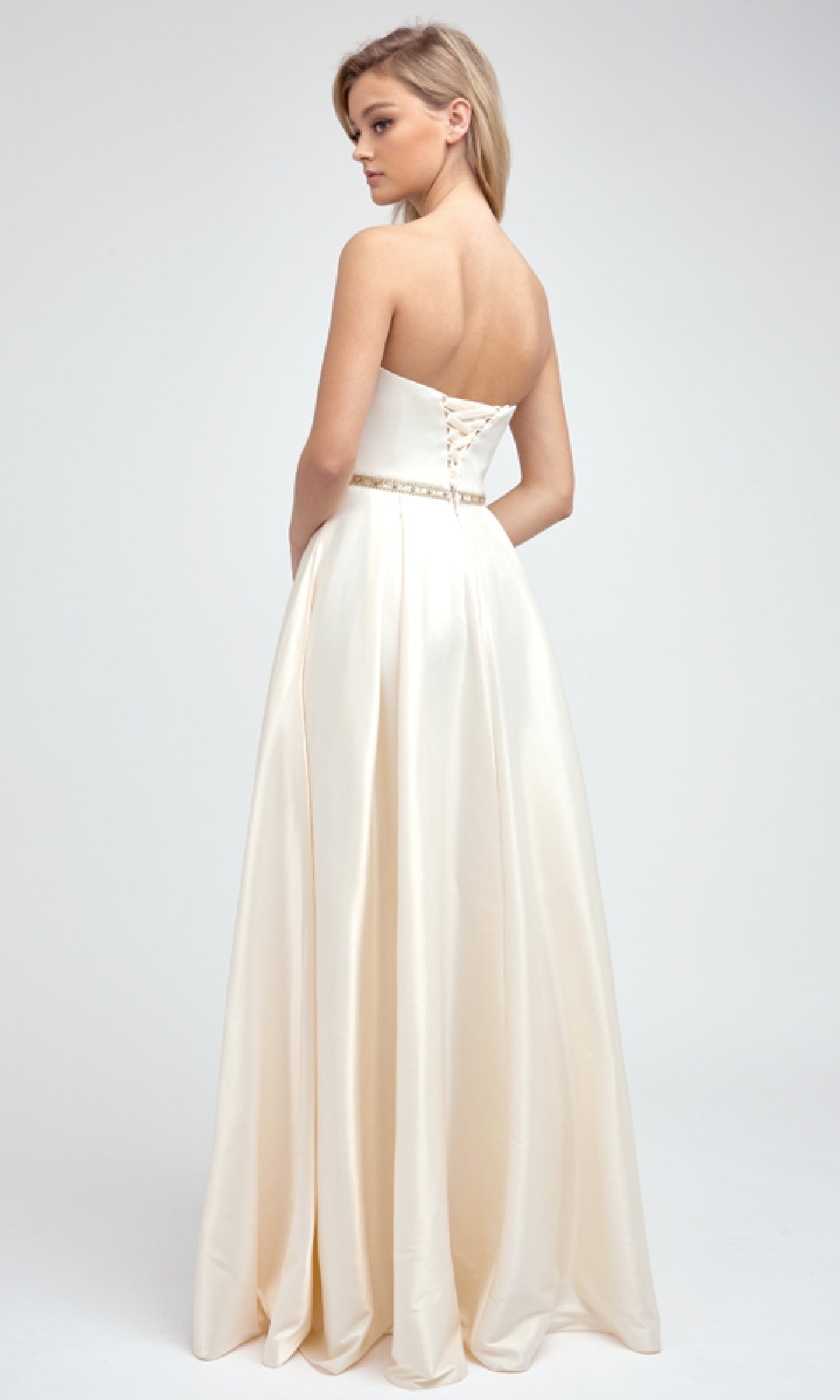A woman stands against a plain light background wearing the Juliet 688, a long ivory satin prom dress with a strapless sweetheart neckline, fitted bodice, lace-up back, and jeweled belt.