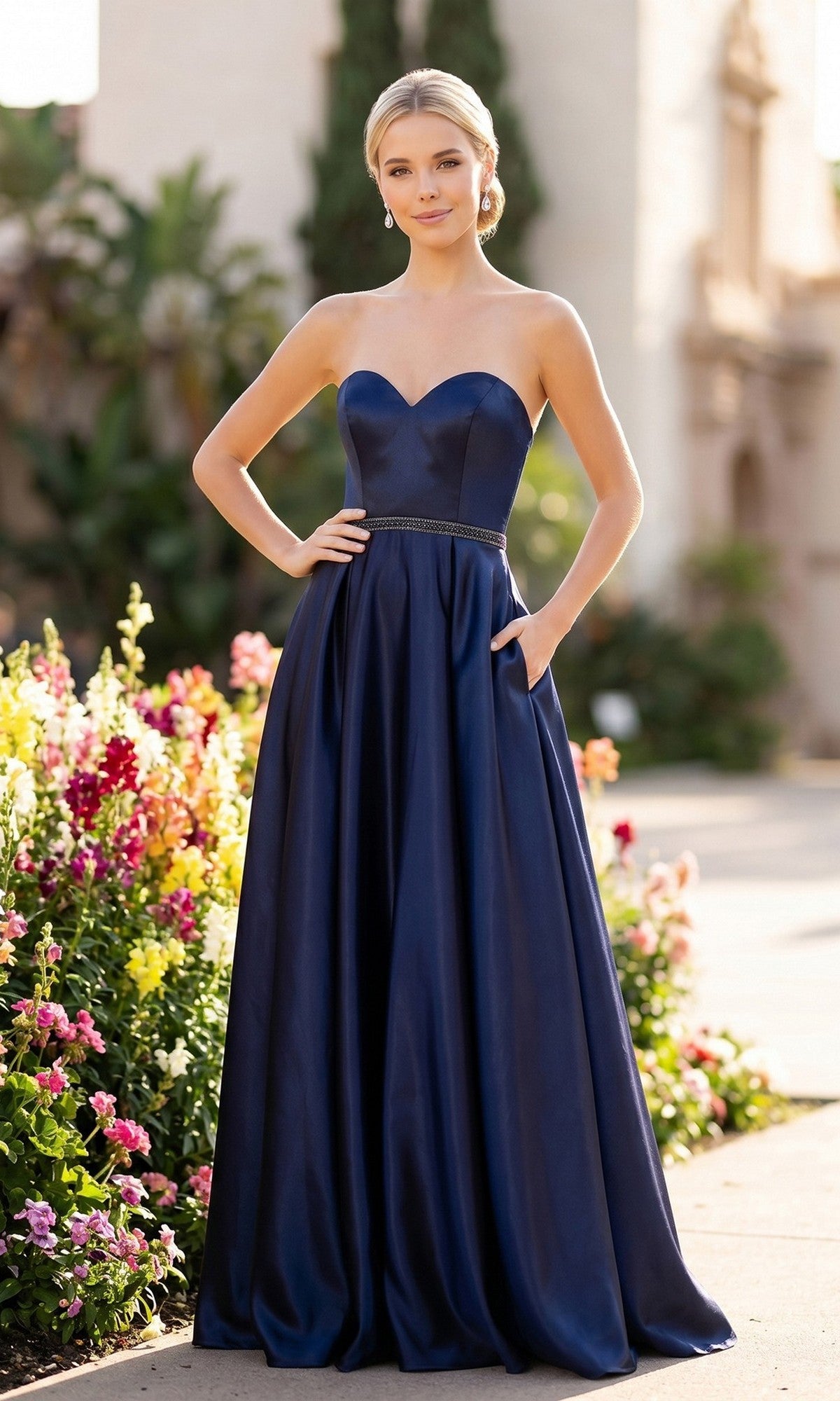 A woman wearing the Juliet 688 long prom dress in navy blue, featuring a strapless fitted bodice and full skirt, stands outdoors near colorful flowers.