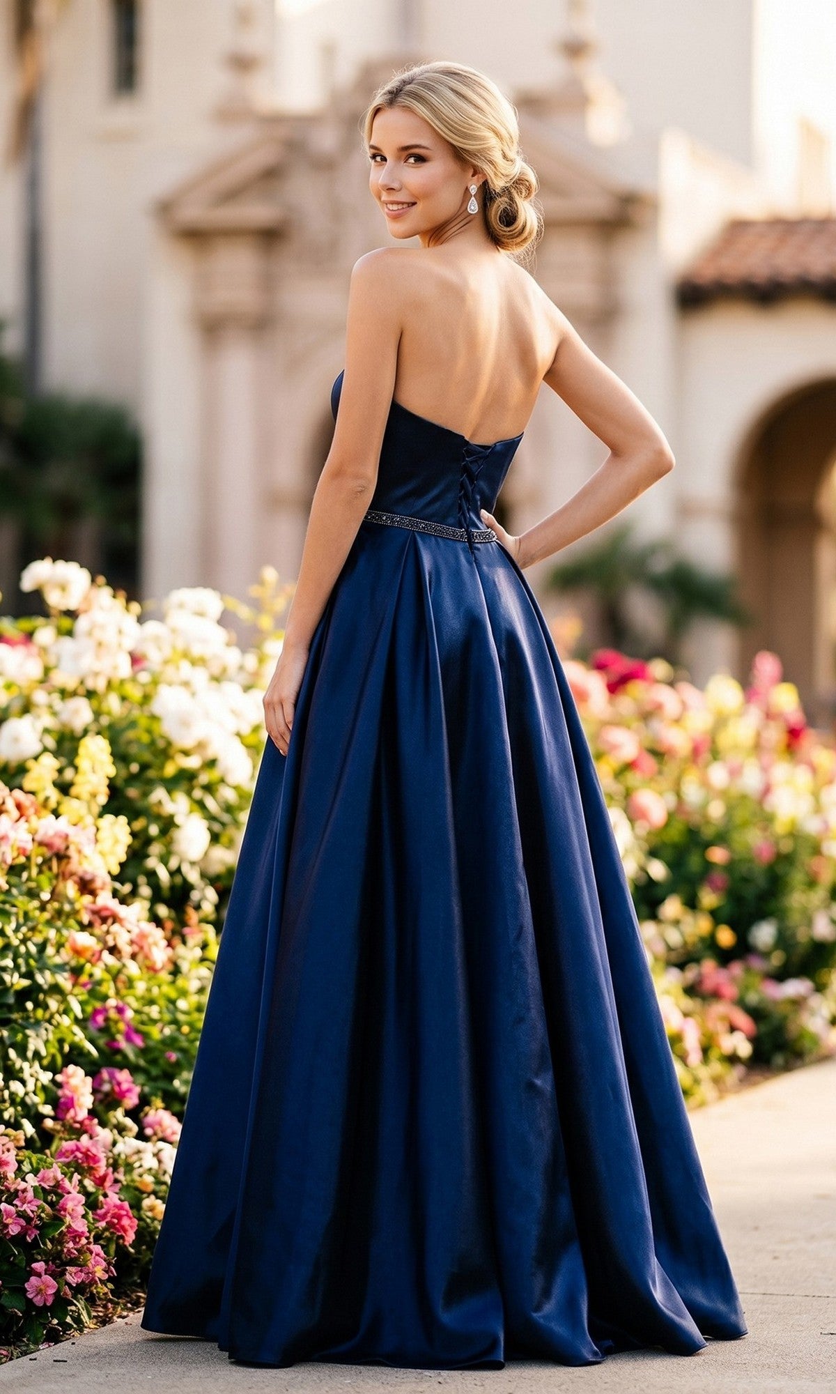 Wearing the Long Prom Dress: Juliet 688—a strapless navy blue gown with a sweetheart neckline—a woman stands outdoors near blooming flowers, softly smiling over her shoulder.