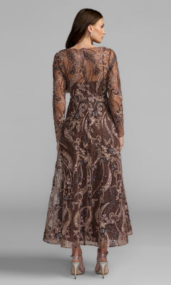 A woman poses with her back to the camera against a plain gray background, wearing the Champagne Midi Party Dress: Mac Duggal 68906—a sheer, long-sleeve embroidered dress in brown and beige tones with a sophisticated midi hemline.