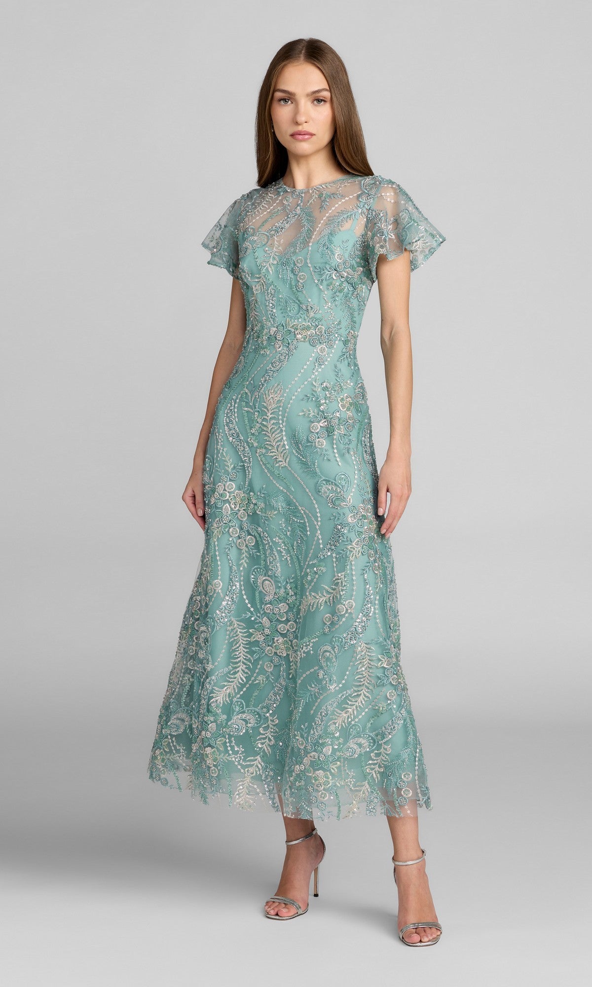 A woman standing in a seafoam tea-length Mac Duggal 68909 party dress with short sleeves, silver floral lace detailing, and delicate beaded embroidery, paired with silver high-heeled sandals against a plain gray background.