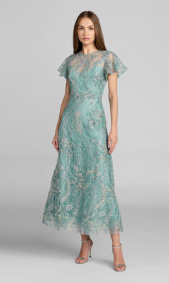 A woman standing in a seafoam tea-length Mac Duggal 68909 party dress with short sleeves, silver floral lace detailing, and delicate beaded embroidery, paired with silver high-heeled sandals against a plain gray background.