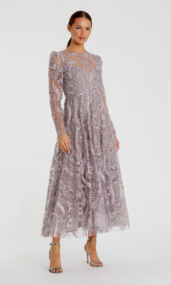A woman stands against a plain background wearing the Mac Duggal 68910 Tea-Length Party Dress in mauve, featuring long sleeves, lace embroidery with floral embellishments, and sheer details. She completes the elegant look—reminiscent of Mac Duggal’s style—with silver high-heeled sandals.