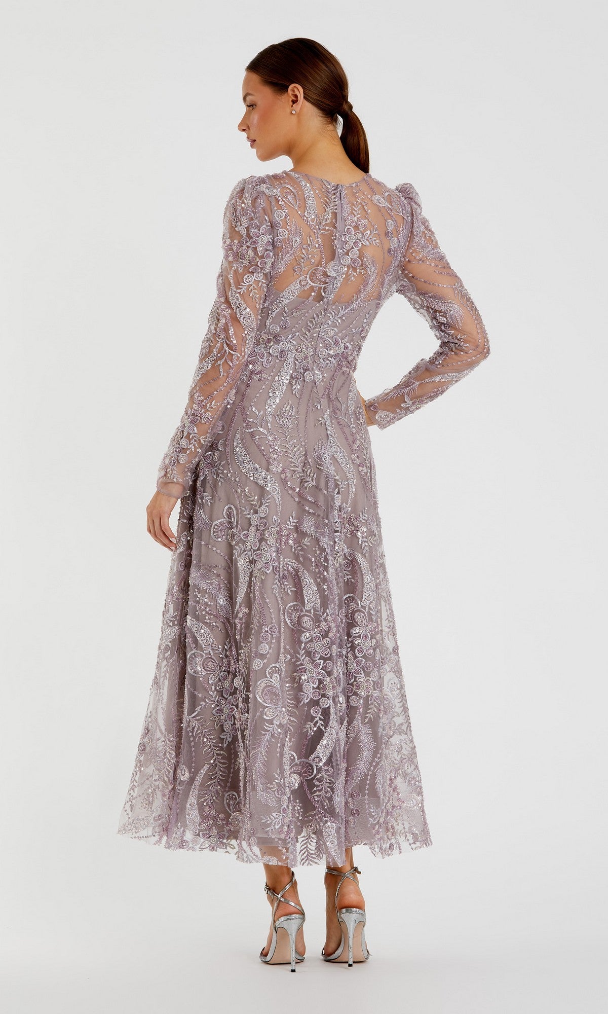 A woman, seen from behind, stands against a plain white background wearing the Mac Duggal 68910 tea-length party dress—a mauve design with long sleeves and floral embroidery—paired with silver heeled sandals.