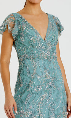 A woman wears the Mac Duggal 68914 Long Formal Dress—a teal gown with short sleeves and a V-neckline, adorned with intricate silver bead and sequin embroidery over delicate lace fabric.