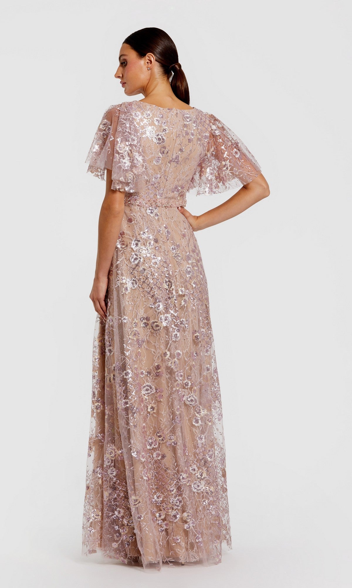 A woman stands with her back turned, wearing the Mac Duggal 68922 long formal dress—a floor-length, short-sleeve gown in light pink lace, beautifully embellished with delicate sequin floral embroidery.