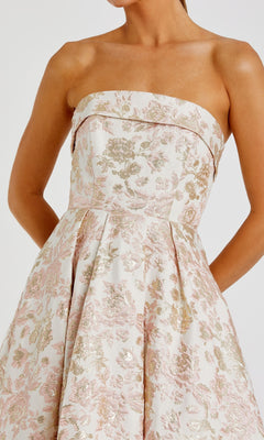 A woman wears the Mac Duggal 68928 long formal dress, a strapless tea-length design in pastel pink and gold floral brocade, standing with her arms relaxed at her sides.