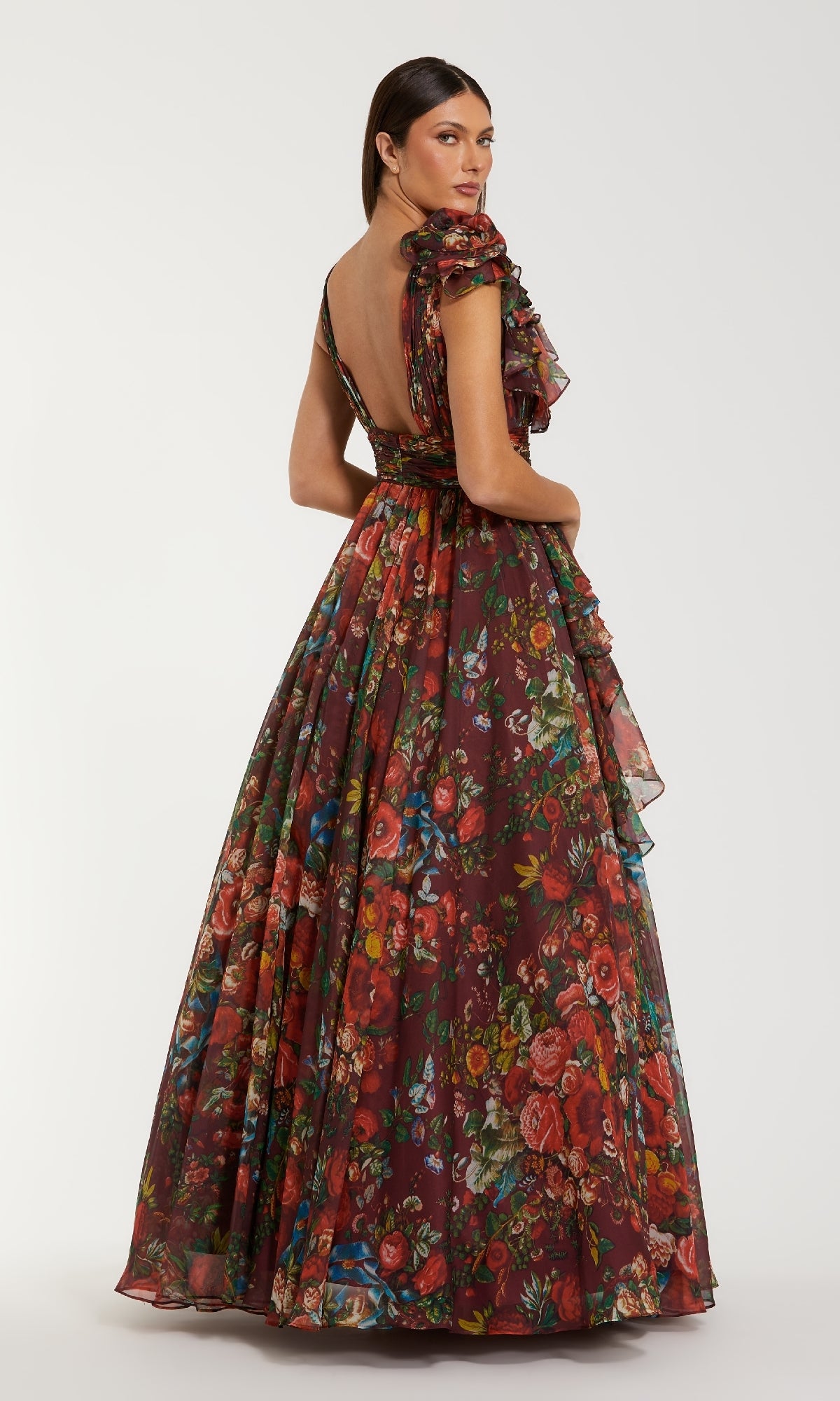 A woman stands facing away from the camera in the Mac Duggal 68947, a long floral-print formal dress with ruffled sleeves and an open back, capturing a sense of timeless elegance.