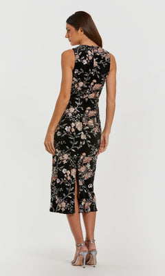 A woman stands in high-heeled sandals, facing away from the camera, wearing the Mac Duggal 68958 midi black wedding guest dress featuring a floral pattern and back slit against a plain background.