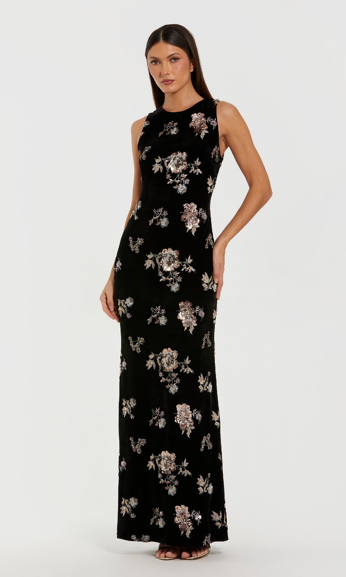 A woman poses in the Mac Duggal 68959 Black Velvet Print Formal Dress, a sleeveless, floor-length gown with a striking floral pattern, set against a plain white background.