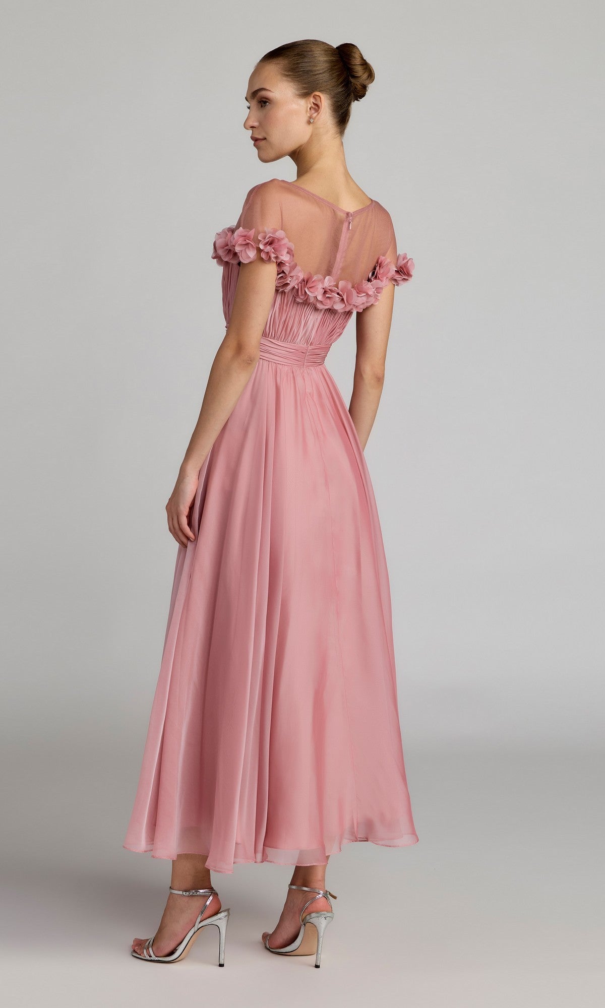 A woman wearing the Mac Duggal 68966 Tea-Length Party Dress—a pink, off-the-shoulder chiffon party dress with floral details—paired with silver heels, stands on a neutral background facing slightly away from the camera.