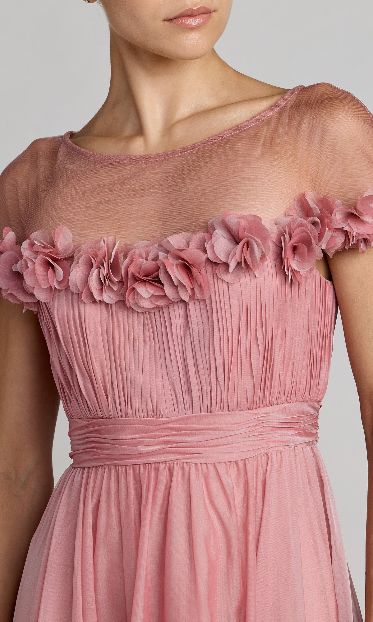 A woman wearing the Tea-Length Party Dress: Mac Duggal 68966, a pink pleated chiffon gown with a sheer neckline and floral appliqués adorning the chest and sleeves—an elegant choice for any special occasion.