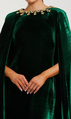A person wearing the Mac Duggal 68977 Green Velvet Caped Formal Dress with gold embellishments around the neckline, hands clasped in front.