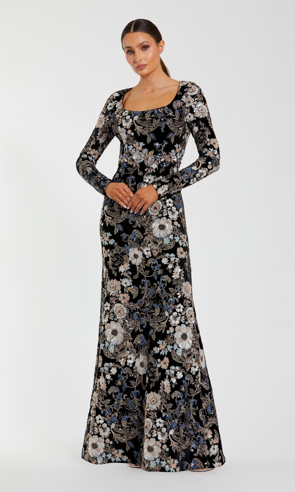 A woman stands facing forward in the Mac Duggal 68978 Black Velvet Print Formal Gown, featuring an intricate embroidered sequin floral print, hands gently clasped at her waist.