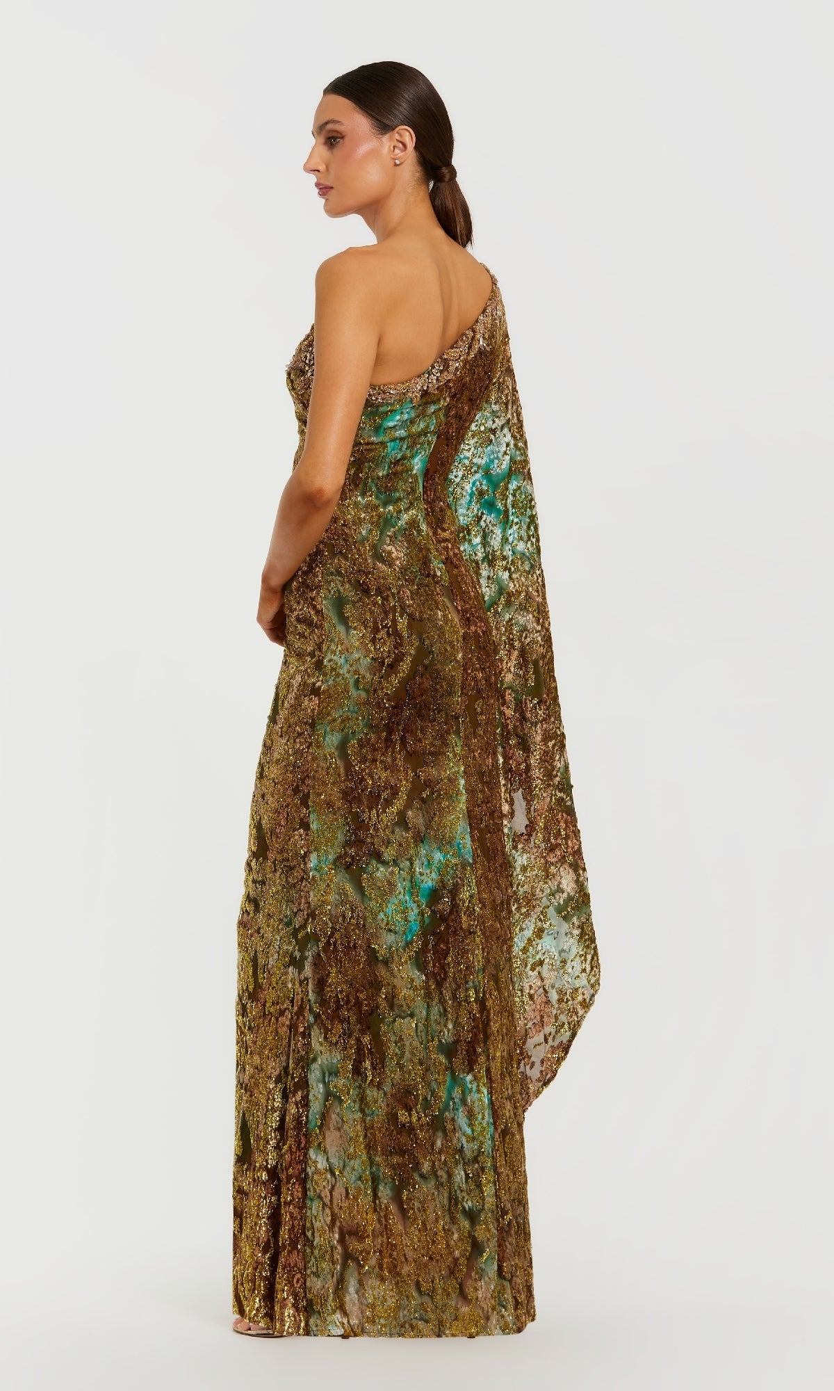 Woman wearing the Mac Duggal 68981 One-Shoulder Gold Formal Gown, featuring a floor-length metallic design with a multicolored pattern, standing sideways against a plain white background.