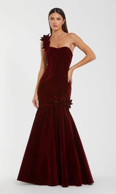 A woman wears the Mac Duggal 68983 One-Shoulder Velvet Formal Dress in deep red, featuring floral accents on the strap and hip, standing against a plain white background.