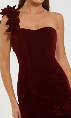 A woman wears the Mac Duggal 68983 one-shoulder velvet formal dress in dark red, featuring a sweetheart neckline and floral embellishments on the shoulder and hip—perfect for formal evenings.